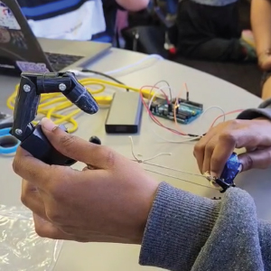 A person demonstrates a 3D-printed mechanical finger connected to strings and powered by a microservo. The setup is wired to an Arduino board on a nearby table, with a laptop and tools in the background